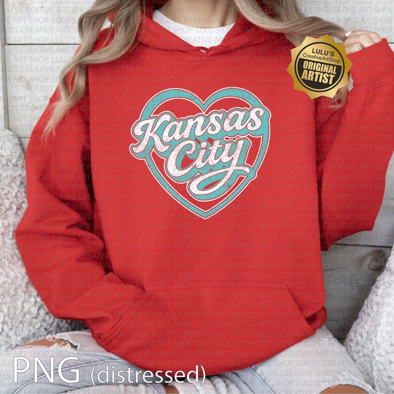 May include: A red hooded sweatshirt featuring a teal heart with the words "Kansas City" in a white, retro font. The sweatshirt has a kangaroo pocket and a drawstring hood. The design includes a "Original Artist" badge.