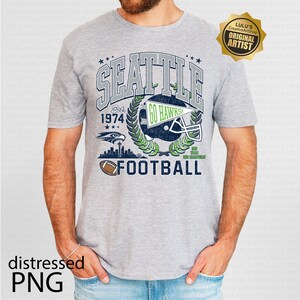 May include: A grey t-shirt with a distressed graphic design. The design includes the word "SEATTLE", a football helmet, and the text "GO HAWKS!". The shirt also features the word "FOOTBALL" and the text "Est. 1974".