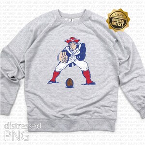 Op de afbeelding: Een lichtgrijs sweatshirt met een verouderde afbeelding van een koloniale soldaat in rood, wit en blauw, die een voetbal vasthoudt. De sweatshirt heeft een ronde hals en lange mouwen. De afbeelding bevat de tekst "distressed PNG".