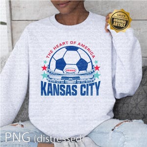 Puede incluir: Sudadera gris claro con un dise&ntilde;o de bal&oacute;n de f&uacute;tbol azul y blanco, el texto "THE HEART OF AMERICA" y "KANSAS CITY". El dise&ntilde;o incluye un gr&aacute;fico de estadio y acentos de estrellas. Un sello dorado "ORIGINAL ARTIST" es visible.