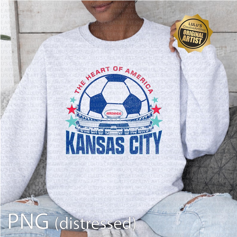 Puede incluir: Sudadera gris claro con un dise&ntilde;o de bal&oacute;n de f&uacute;tbol azul y blanco, el texto "THE HEART OF AMERICA" y "KANSAS CITY". El dise&ntilde;o incluye un gr&aacute;fico de estadio y acentos de estrellas. Un sello dorado "ORIGINAL ARTIST" es visible.