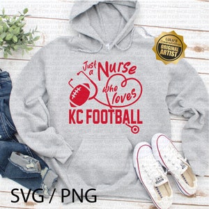 K&ouml;nnte beinhalten: Ein grauer Kapuzenpullover mit dem roten Text "Just a Nurse Who Loves KC FOOTBALL". Das Design umfasst einen Football, ein Stethoskop und ein Herz. Wei&szlig;e Converse-Sneaker und Jeans sind ebenfalls im Bild.