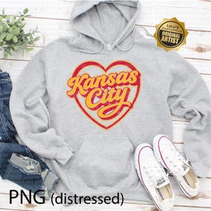May include: A light gray hoodie with a front pocket and a hood. The hoodie features a distressed design of a red heart with the words "Kansas City" in yellow script. White sneakers are placed near the hoodie. The image also includes the text "PNG (distressed)."