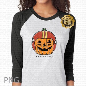 May include: White and black raglan-style shirt with a graphic of an orange pumpkin wearing a football helmet. The pumpkin has a carved face and the text "Kansas City" is below the pumpkin.