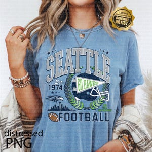 May include: Pale blue t-shirt with a Seattle football graphic. The design includes the words "SEATTLE", "GO HAWKS!", a football helmet, and the year "1974". The shirt has a distressed PNG graphic.