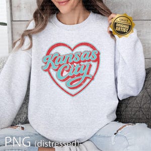May include: A light gray sweatshirt featuring a distressed red heart with the words "Kansas City" in turquoise script. The sweatshirt has a crew neck and long sleeves. The image also includes a gold sticker that says "Original Artist".