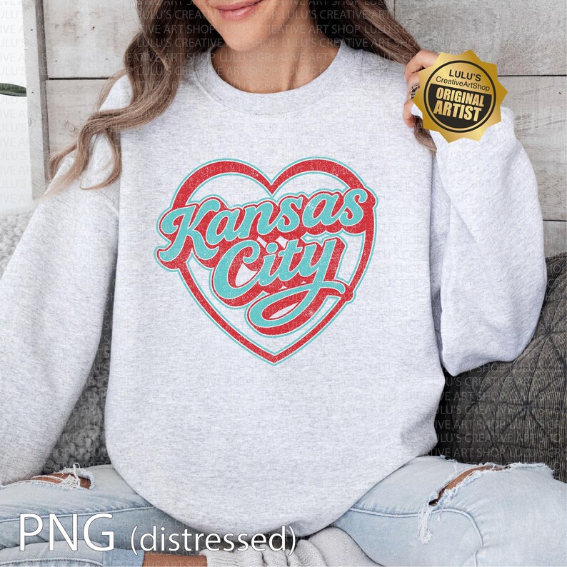 May include: A light gray sweatshirt featuring a distressed red heart with the words "Kansas City" in turquoise script. The sweatshirt has a crew neck and long sleeves. The image also includes a gold sticker that says "Original Artist".