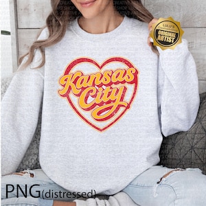 May include: A light gray sweatshirt featuring a distressed red heart with the words "Kansas City" in yellow script. The sweatshirt has a crew neck and long sleeves. The image also includes a gold "Original Artist" sticker.