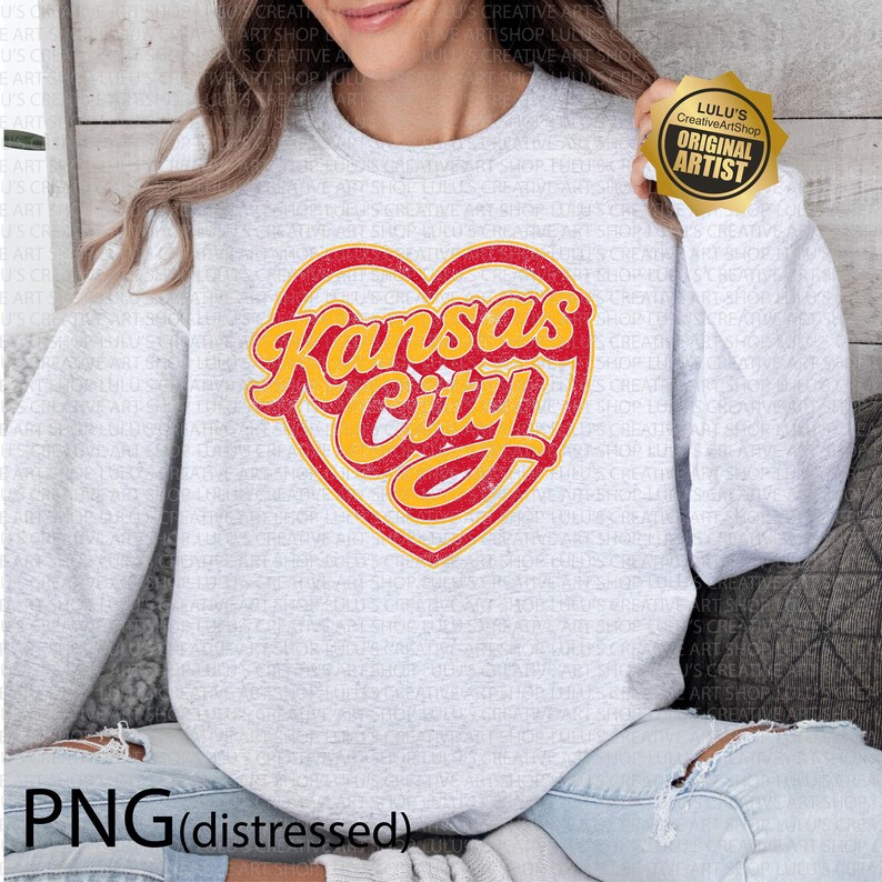 May include: A light gray sweatshirt featuring a distressed red heart with the words "Kansas City" in yellow script. The sweatshirt has a crew neck and long sleeves. The image also includes a gold "Original Artist" sticker.