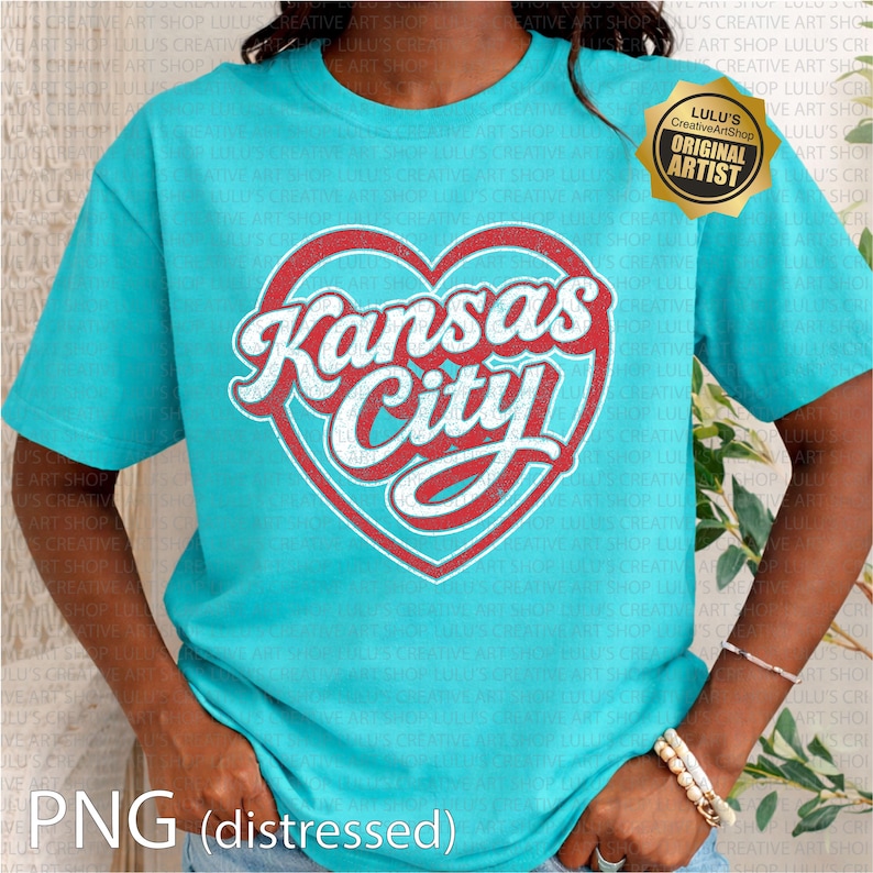 May include: A turquoise t-shirt featuring a red and white heart-shaped design with the words "Kansas City" in a vintage style. The shirt has short sleeves and a crew neck. A gold-colored "Original Artist" seal is visible.