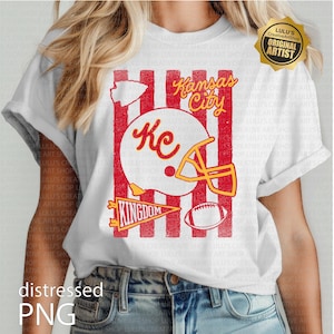 May include: A white t-shirt featuring a graphic print with vertical red stripes, a white football helmet with the letters KC, a yellow facemask, a pennant with the word KINGDOM, a football, and the text Kansas City in yellow cursive script.