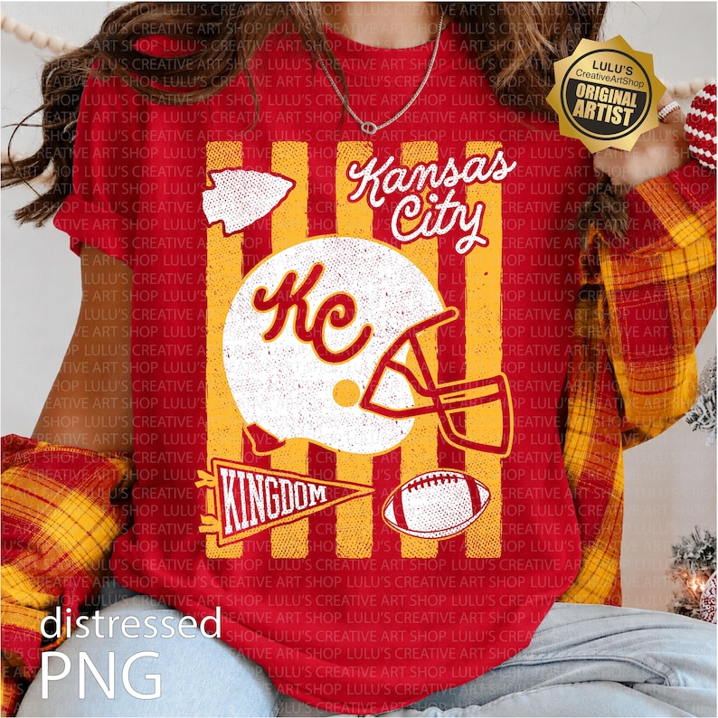 May include: A red t-shirt featuring a graphic print with vertical yellow stripes. The design includes a white football helmet with the letters KC, a football, an arrowhead, a pennant with the word KINGDOM, and the text Kansas City.