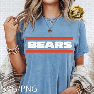 May include: Light blue t-shirt with the word "BEARS" in white block letters, framed by two horizontal orange stripes. The shirt has short sleeves and a relaxed fit. A gold-colored "Original Artist" sticker is visible.