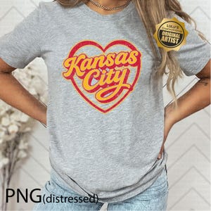 May include: A heather gray t-shirt featuring a red heart with the words "Kansas City" in yellow script. The shirt has short sleeves and a crew neck. The text "PNG (distressed)" is at the bottom.