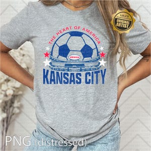 Puede incluir: Una camiseta gris con un dise&ntilde;o de bal&oacute;n de f&uacute;tbol azul y blanco, con el texto "THE HEART OF AMERICA" y "KANSAS CITY". El dise&ntilde;o incluye un gr&aacute;fico de estadio y acentos de estrellas. La camiseta tambi&eacute;n tiene el texto "ARROWHEAD".