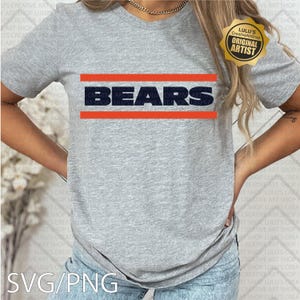 May include: A heather gray t-shirt with the word "BEARS" in navy blue block letters, framed by two orange horizontal lines. The shirt also has a gold-colored "ORIGINAL ARTIST" seal.