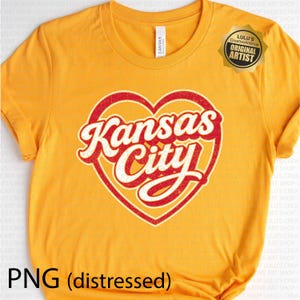 May include: Mustard yellow t-shirt with a distressed red and white heart-shaped graphic that reads "Kansas City". The shirt has a crew neck and short sleeves. A gold sticker with text is on the upper right.