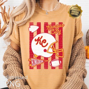 May include: A mustard yellow t-shirt featuring a graphic print with red vertical stripes, a white football helmet with the letters Kc, a white arrowhead, a yellow pennant with the word KINGDOM, a football, and the text Kansas City in yellow.
