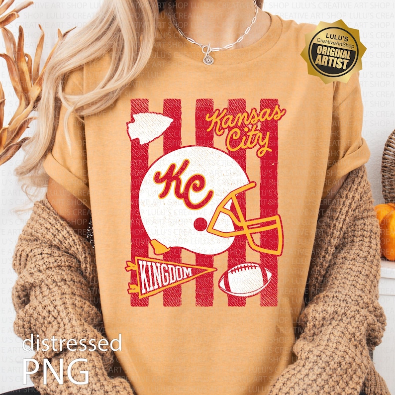 May include: A mustard yellow t-shirt featuring a graphic print with red vertical stripes, a white football helmet with the letters Kc, a white arrowhead, a yellow pennant with the word KINGDOM, a football, and the text Kansas City in yellow.