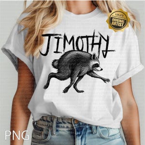 May include: A white short-sleeve t-shirt featuring a black ink-style illustration of a raccoon in a running pose. Above the raccoon, the text JIMOTHY is printed in a bold, jagged, hand-drawn black font. A gold-colored badge is in the top right corner.