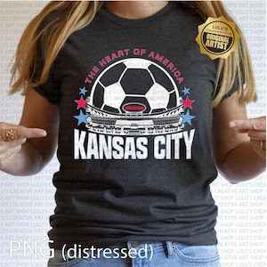 Puede incluir: Camiseta gris oscuro con un dise&ntilde;o de bal&oacute;n de f&uacute;tbol, el texto "The Heart of America" y "Kansas City". El dise&ntilde;o incluye un gr&aacute;fico de estadio y acentos de estrellas rojas y azules. Una insignia dorada de "Original Artist" es visible.
