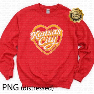 May include: Red crewneck sweatshirt featuring a heart-shaped design with "Kansas City" in a stylized font. The design is in yellow and white. The sweatshirt also has a "Lulu's Creative Art Shop" logo.