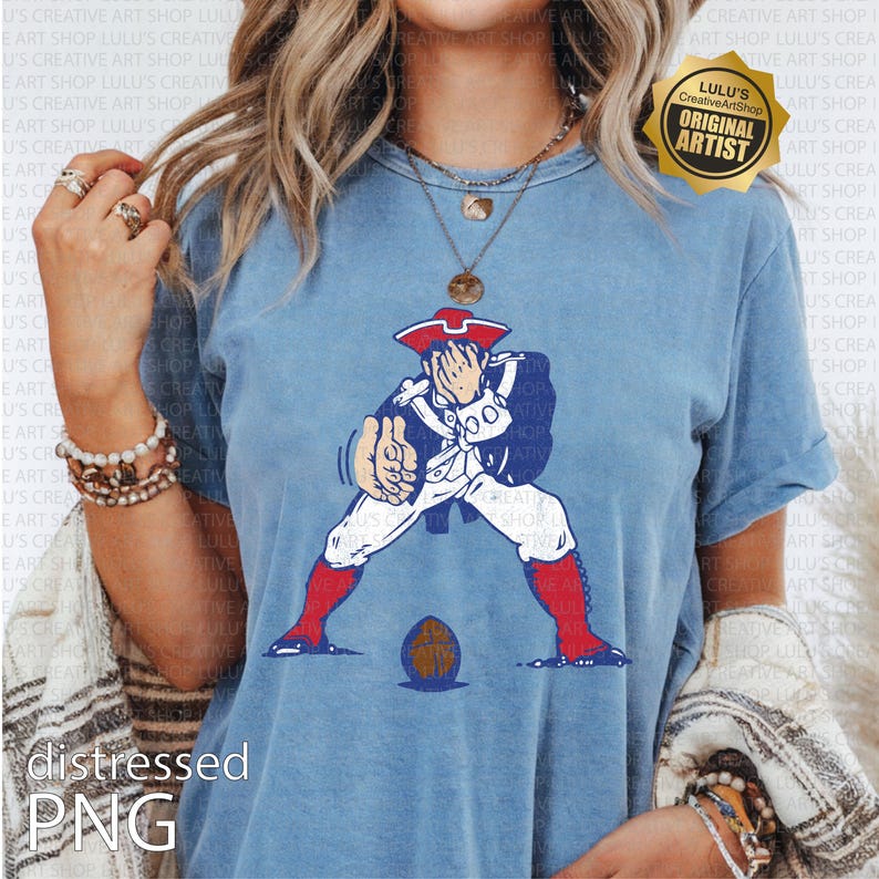 Op de afbeelding: Lichtblauw T-shirt met een cartoonillustratie van een koloniale soldaat in een patriottische pose. De soldaat draagt een witte broek, een blauwe jas en een rode hoed en laarzen. Een voetbal ligt aan de voeten van de soldaat. Het shirt heeft de tekst "distressed PNG".