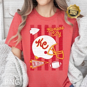 May include: A red short-sleeve t-shirt featuring a graphic print with vertical stripes, a white football helmet with the letters KC, a football, a pennant with the word KINGDOM, an arrowhead shape, and the text Kansas City in yellow script.