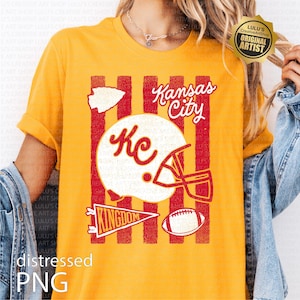 May include: A mustard yellow t-shirt featuring a graphic print with red vertical stripes, a white football helmet with the letters KC, a white arrowhead, a red pennant with the word KINGDOM, a football, and the text Kansas City in white script.