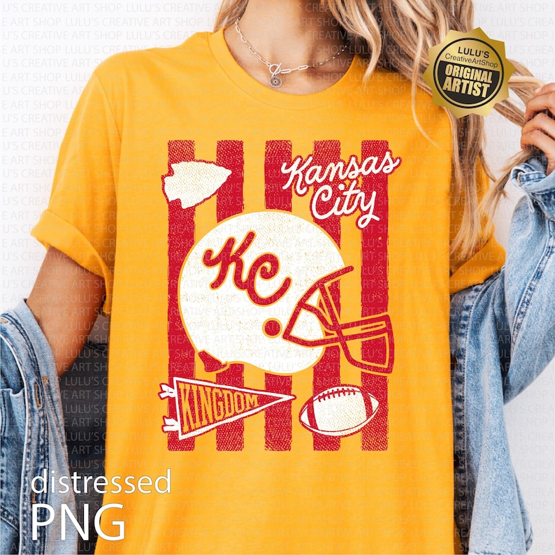 May include: A mustard yellow t-shirt featuring a graphic print with red vertical stripes, a white football helmet with the letters KC, a white arrowhead, a red pennant with the word KINGDOM, a football, and the text Kansas City in white script.