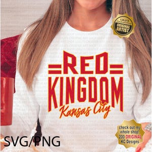 May include: A white t-shirt with a red and yellow graphic that says "=RED= KINGDOM Kansas City". The graphic is in a bold, blocky font. The t-shirt is being worn by a person.
