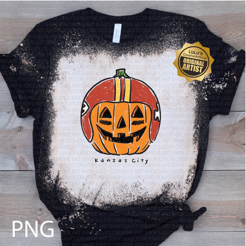 May include: Black t-shirt with a distressed bleach effect, featuring a hand-drawn pumpkin wearing a football helmet. The pumpkin has a carved face and the text "Kansas City" below. A gold seal reads "Original Artist".