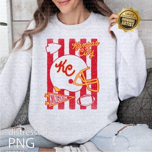 May include: A light gray crewneck sweatshirt featuring a red and white graphic print. The design includes vertical red stripes, a white football helmet with the text KC, the words Kansas City, a pennant with the text KINGDOM, and a football.