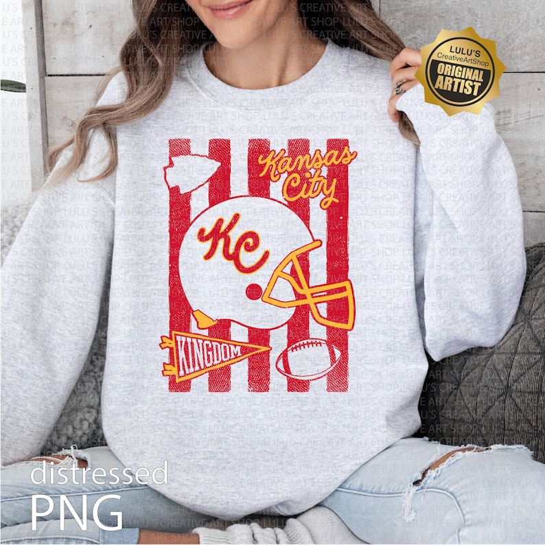 May include: A light gray crewneck sweatshirt featuring a red and white graphic print. The design includes vertical red stripes, a white football helmet with the text KC, the words Kansas City, a pennant with the text KINGDOM, and a football.