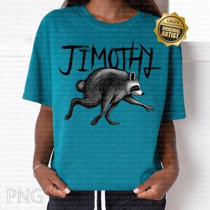 May include: A teal short-sleeved t-shirt featuring a black ink-style illustration of a raccoon in a running pose. Above the raccoon, the name "JIMOTHY" is printed in a jagged, hand-drawn black font. A gold badge in the corner reads "LULU'S ORIGINAL ARTIST".