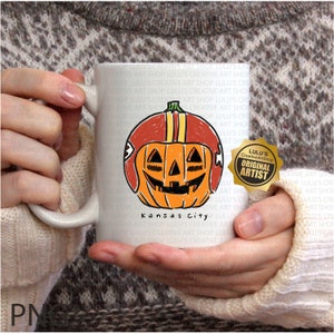 May include: White ceramic mug featuring a hand-drawn pumpkin wearing a football helmet. The pumpkin has a carved face and is colored orange, with a red helmet and yellow stripe. The text "Kansas City" is below the pumpkin.