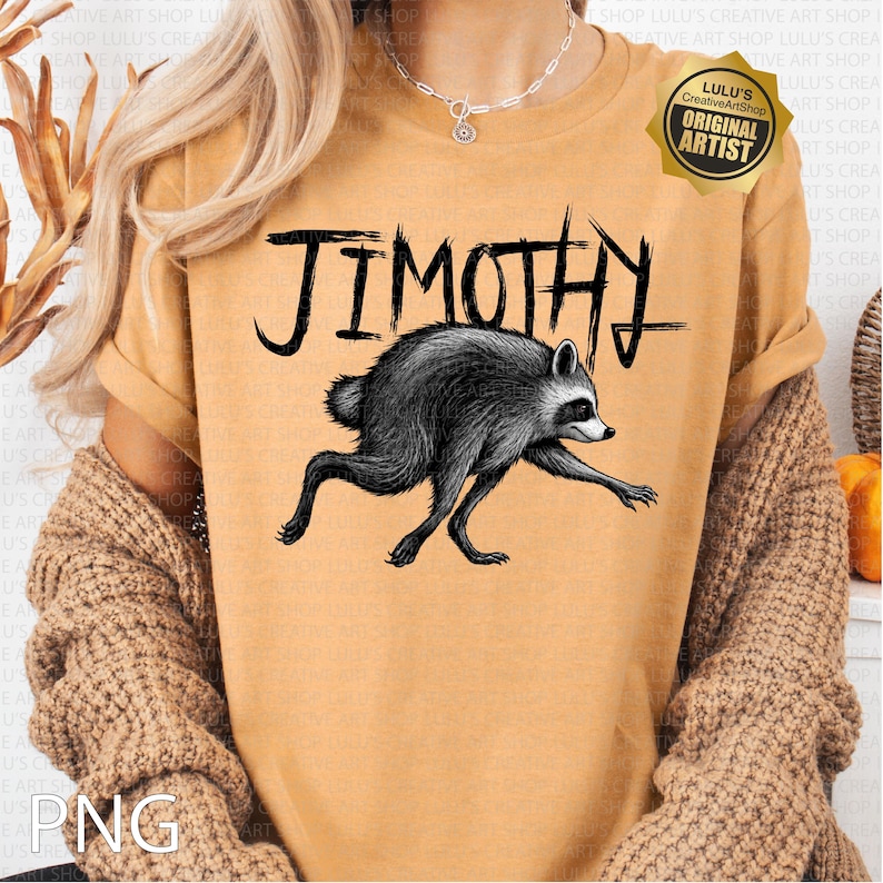 May include: A mustard yellow t-shirt featuring a black graphic of a raccoon with the text "JIMOTHY" written in a stylized, jagged font above the animal. The shirt has short sleeves and a crew neckline, with a gold badge in the top right corner.