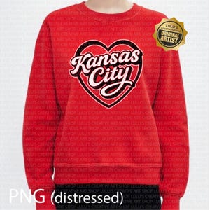 May include: Red crewneck sweatshirt featuring a heart-shaped design with the words "Kansas City" in white script and black outline. The sweatshirt has long sleeves and a classic fit. The image also includes a gold-colored badge with the text "Original Artist".