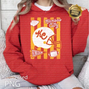 May include: A red hooded sweatshirt featuring a graphic print with yellow vertical stripes. The design includes a white football helmet with the letters KC, a football, a pennant with the word KINGDOM, and the text Kansas City in white script.