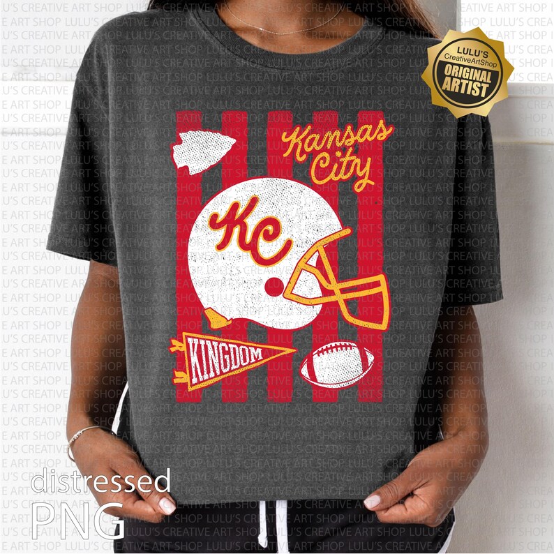 May include: A dark gray t-shirt featuring a graphic print with red vertical stripes, a white football helmet with the letters KC, a football, an arrowhead, and a pennant with the word KINGDOM. The text Kansas City is printed in yellow.