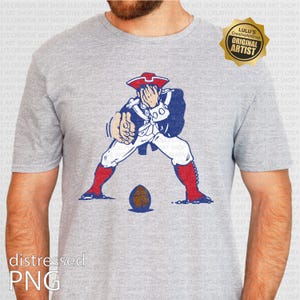 Op de afbeelding: Grijze t-shirt met een verweerde afbeelding van een koloniale figuur in een steek, witte jas en rode laarzen, klaar om een bruine voetbal te trappen. Het ontwerp bevat de tekst "distressed PNG".