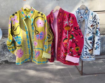 Puede incluir: Tres chaquetas de manga larga colgadas en perchas de madera. La chaqueta de la izquierda es de color amarillo mostaza con bordados florales rosas y azules. La central es magenta con bordados florales multicolores. La de la derecha es blanca con bordados florales azules y marrones.