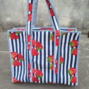 May include: A rectangular tote bag with a white and blue striped pattern, adorned with red strawberry illustrations. The bag has matching striped handles and trim, creating a cohesive design. The bag is made of fabric.
