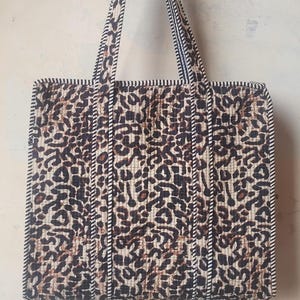May include: A quilted tote bag with a leopard print pattern in shades of brown and tan. The bag has sturdy handles and is trimmed with a black and white striped border. The bag is designed for carrying items.