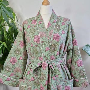 Könnte beinhalten: Grüner Bademantel mit rosa Blumenmuster und weißer Paspelierung. Der Bademantel wird auf einer Schaufensterpuppe präsentiert und hat einen passenden Bindegürtel und Taschen. Der Bademantel ist aus einem weichen, leichten Stoff gefertigt.