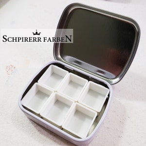 May include: A metal watercolor paint palette with six empty pans. The palette is open and the lid is silver. The brand name "SCHPIRERR FARBEN" is printed on the lid.