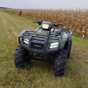 Op de afbeelding: Een olijfgroen Honda terreinvoertuig met een zwarte bagagedrager aan de voorzijde, een centrale koplamp en twee heldere LED-koplampen geïntegreerd in de grille. Het voertuig heeft grote banden met een diep profiel en een robuust frame.
