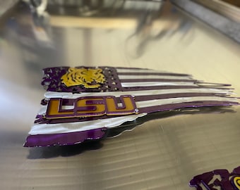 May include: A purple, white and gold metal American flag with a distressed look. The flag has a tiger head cutout and the letters "LSU" in gold and purple.