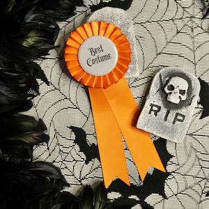 May include: Orange and white Halloween ribbon with the text "Best Costume" on a white background. The ribbon is on a black and white spiderweb patterned fabric with a white tombstone with a skull and the text "RIP" on it.