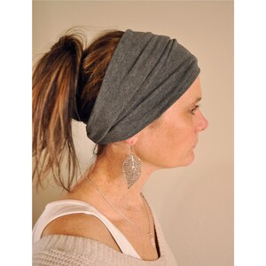 May include: A gray headband worn around the forehead. The headband is made of a soft fabric. The person is also wearing a silver leaf-shaped earring and a silver necklace. The hair is pulled back in a ponytail.